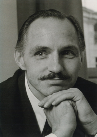 A black and white photo of a 40-ish man with black hair and a moustache, looking a little like Glark Gable. He is resting his chin on his folded hands and wearing a dark suit, white shirt and a tie.