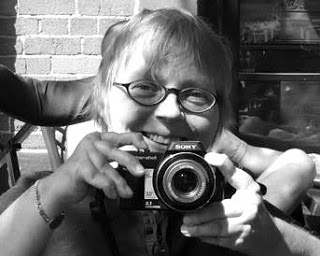 Black-and-white photo of younger version of me, a white woman glasses and short hair pointing a camera at the photographer and smiling. My hands and fingers show obvious RA deformities.