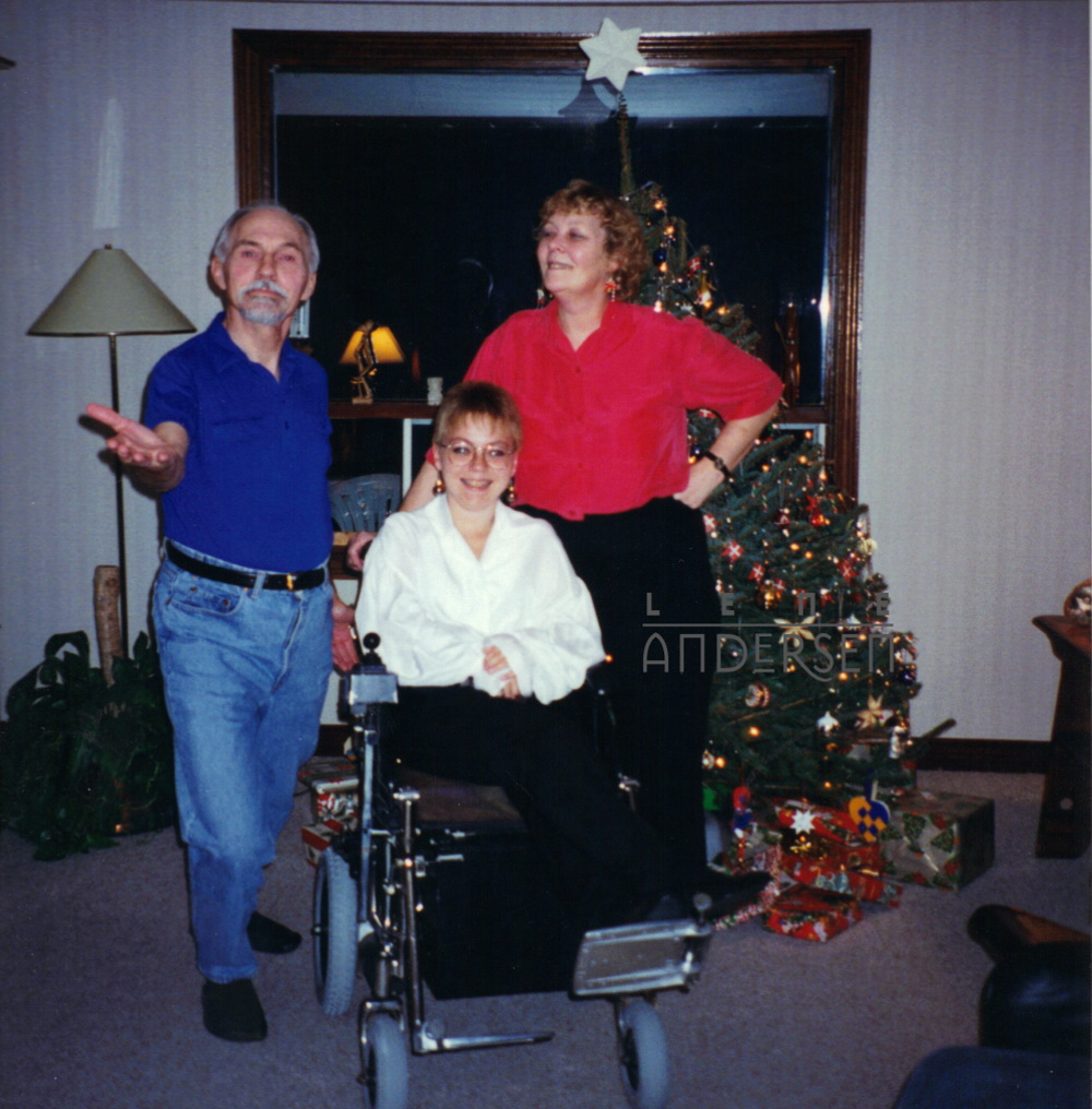 A young white woman with short hair and glasses (me() is sitting in a wheelchair in front of a Christmas tree. Her mother is standing to the right behind the chair and her father to the left. He's doing a fancy lunge, hand outstretched as if saying "ta-da". Both women are smiling.