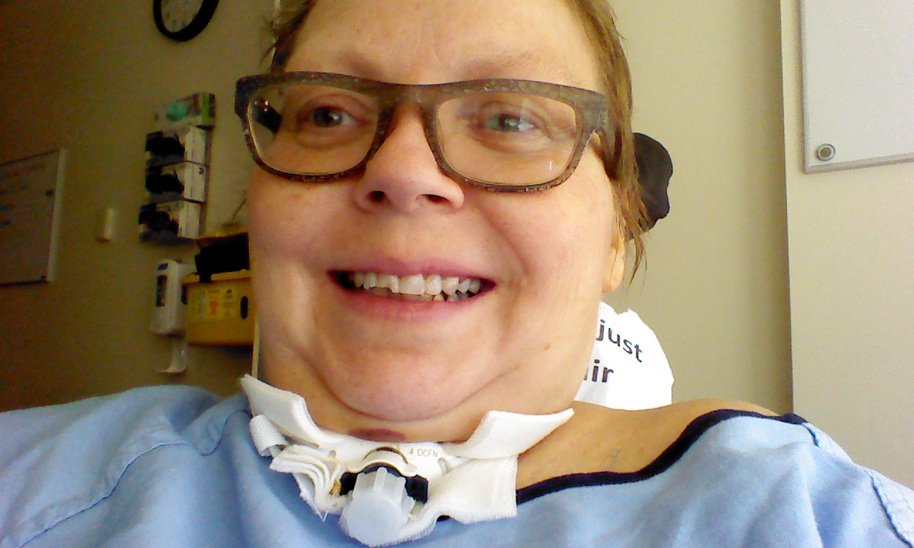 A closeup of me, a white woman with glasses and short hair, sitting in my wheelchair. I have the trach, but it's capped. I am smiling widely.
