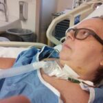 A photo of me, a white woman with shirt hair and glasses) in an ICU bed. The view is from the side and you can clearly see the tracheostomy